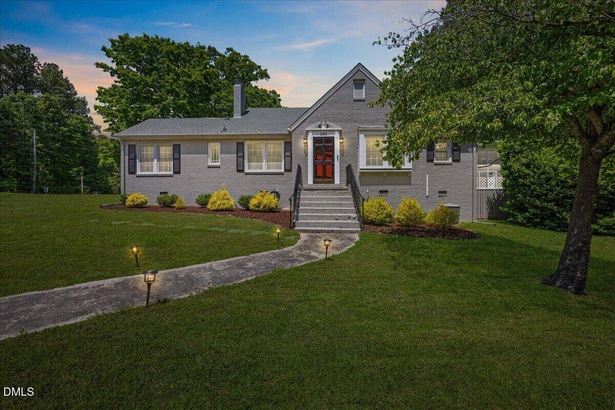 Charming gray single-story house with a red door, steps leading to the entrance, a curved path with garden lights, and a well-maintained lawn with trees and shrubs, set against a partly cloudy sky at dusk.