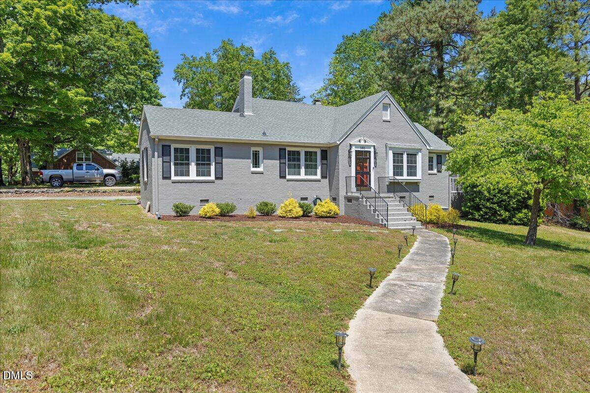 A light gray house with white trim sits on a grassy, slightly sloped yard. A curved sidewalk leads to the front steps, and small shrubs line the house. Tall trees and a parked car are visible in the background.