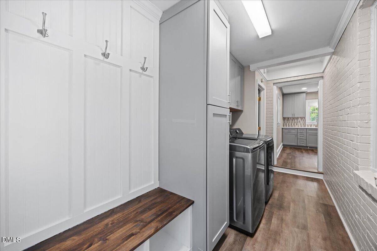 A laundry room with a wooden bench and hooks on the left, gray cabinets, a washer and dryer in the center, and a view into another room with large windows.