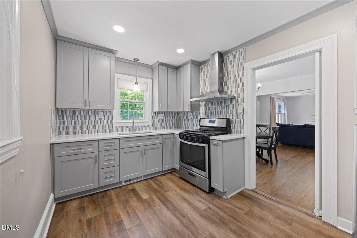 Modern kitchen with gray cabinets, patterned backsplash, stainless steel gas stove, and wooden floors. A window above the sink lets in natural light. An open doorway leads to a dining area and living room with dark blue sofa.