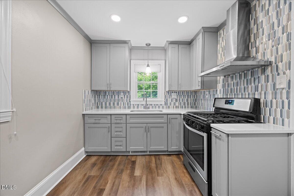 Modern kitchen with gray cabinets, stainless steel gas stove and hood, wood floors, and hexagonal tile backsplash. A window above the sink lets in natural light, brightening the space.
