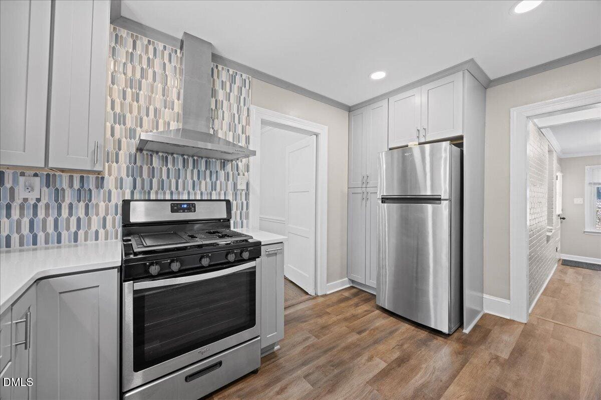 Modern kitchen with light gray cabinets, stainless steel appliances including a stove and refrigerator, patterned tile backsplash, wood floor, and recessed lighting. Two white doors and a hallway are visible.