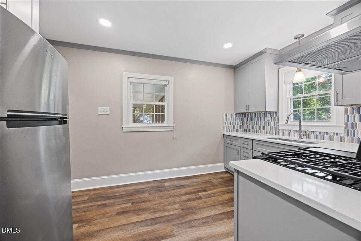Modern kitchen with light gray cabinets, stainless steel refrigerator, gas stove, colorful tile backsplash, white countertops, wood-style flooring, and two windows letting in natural light.