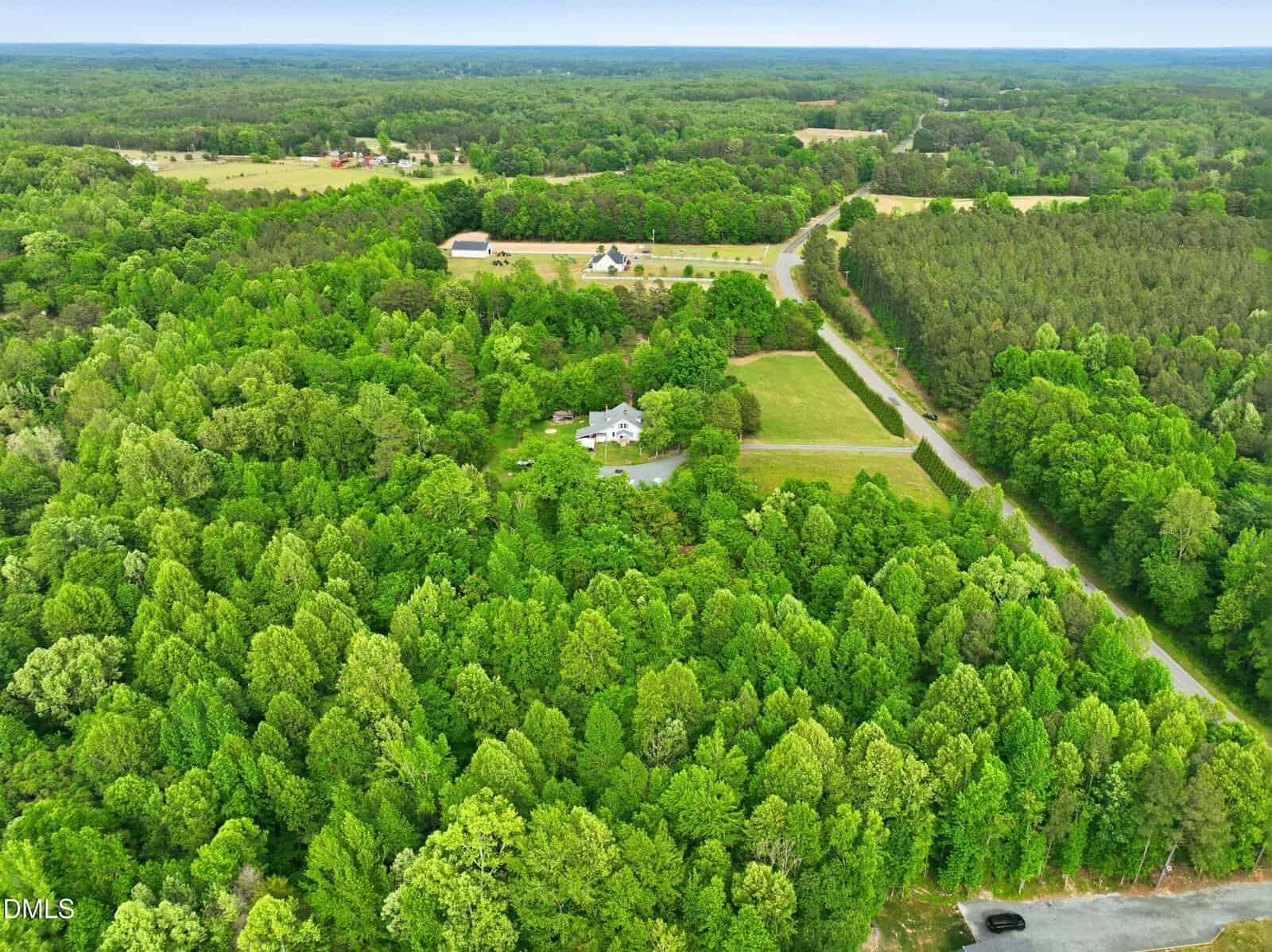 Aerial view of a white house surrounded by dense green forest and open grassy areas, with a pond nearby and roads curving through the landscape. Additional homes and buildings are visible in the background.