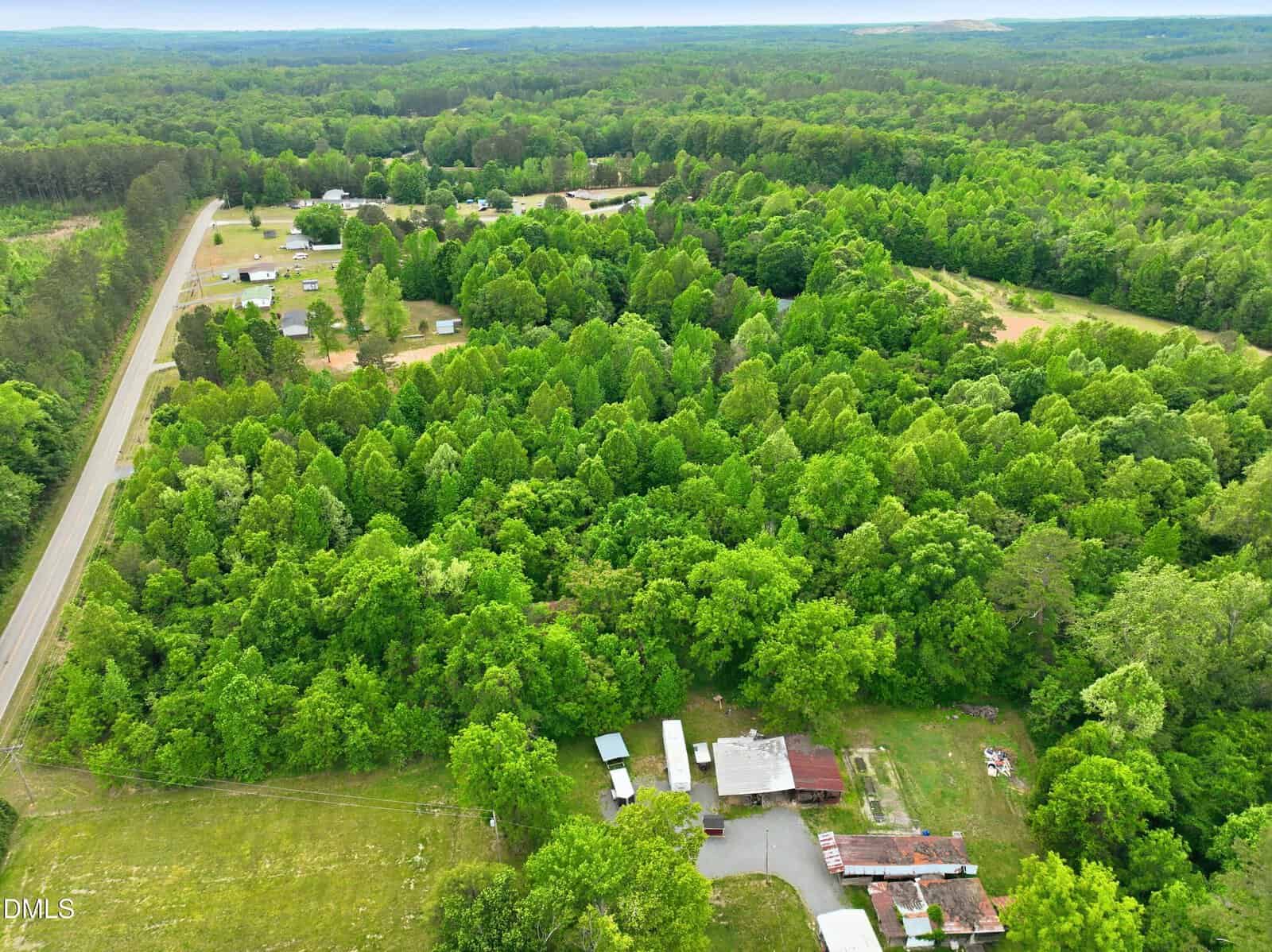 Aerial view of a rural property with several buildings surrounded by dense green trees, a road running along the left edge, and multiple homes visible in the background amid forested areas.