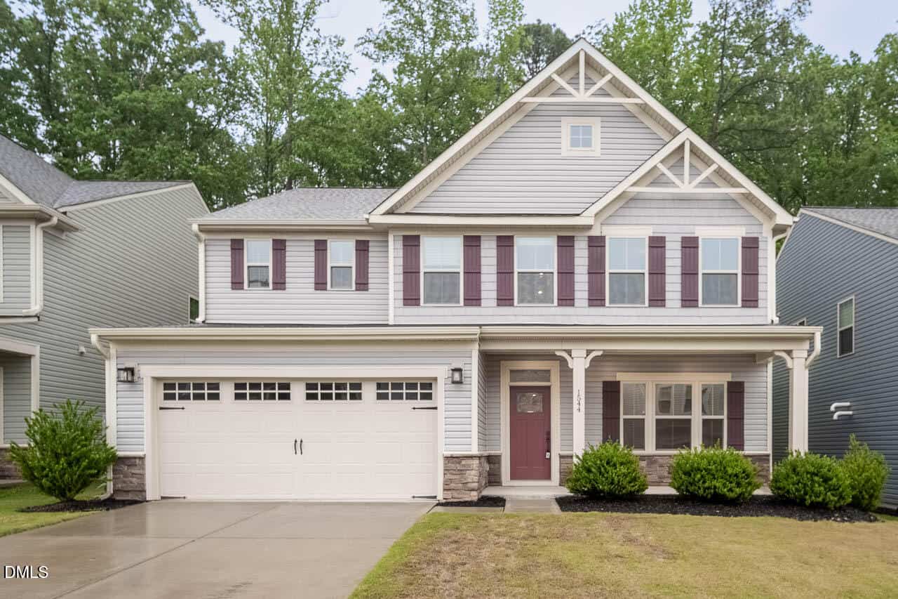 Two-story suburban house with gray siding, maroon shutters, a covered front porch, double garage, and manicured bushes, set against a backdrop of trees and neighboring homes.