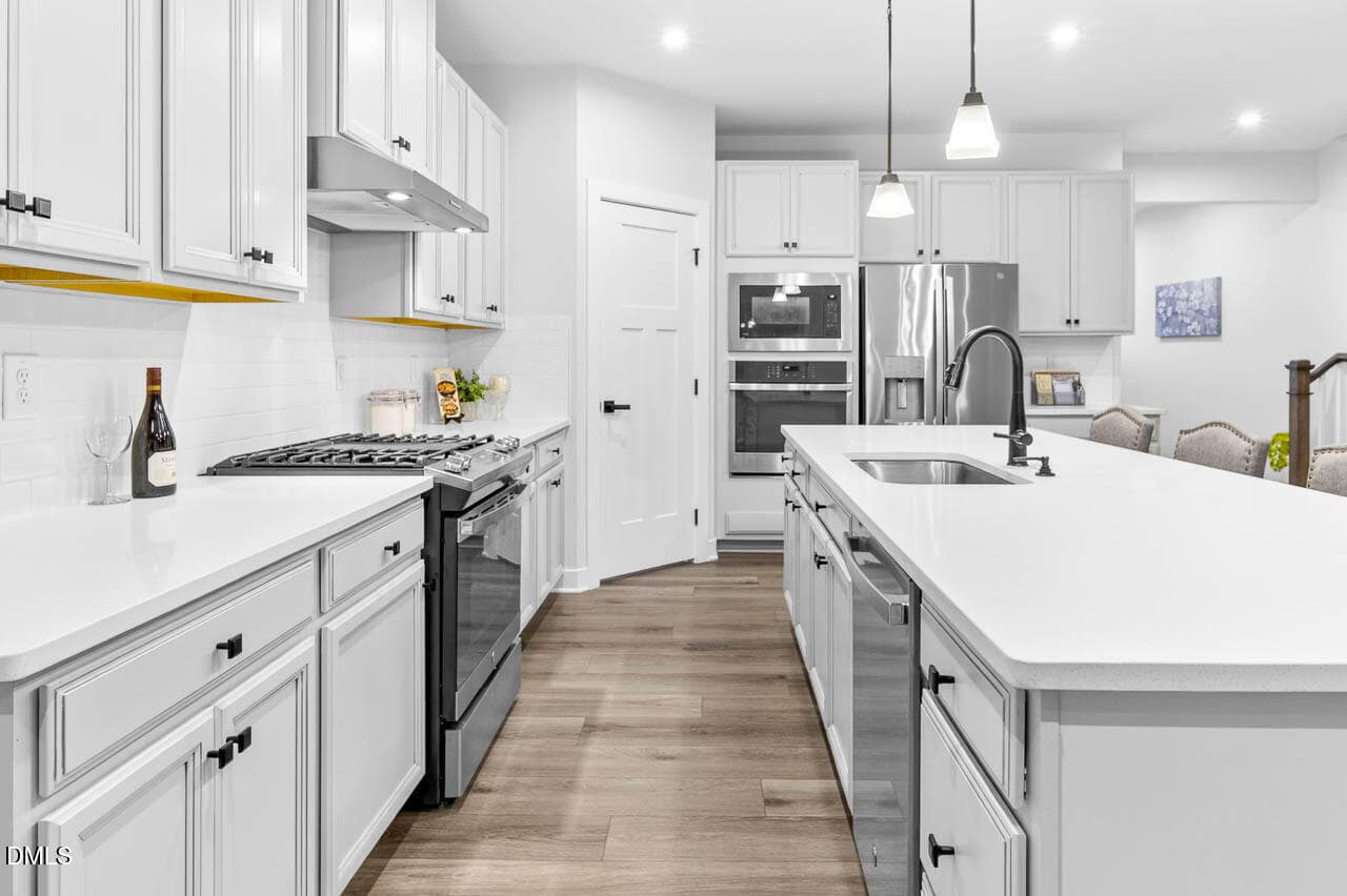 Modern kitchen with white cabinets, stainless steel appliances, a center island with a sink and faucet, pendant lights, and light wood flooring. A bottle of wine and fruit sit on the counter. Dining area visible in the background.