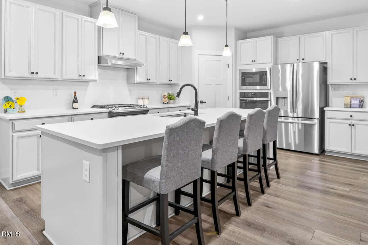 Modern kitchen with white cabinets, stainless steel appliances, a large island with a sink, and four gray upholstered barstools. Pendant lights hang above the island, and the floor is light wood.