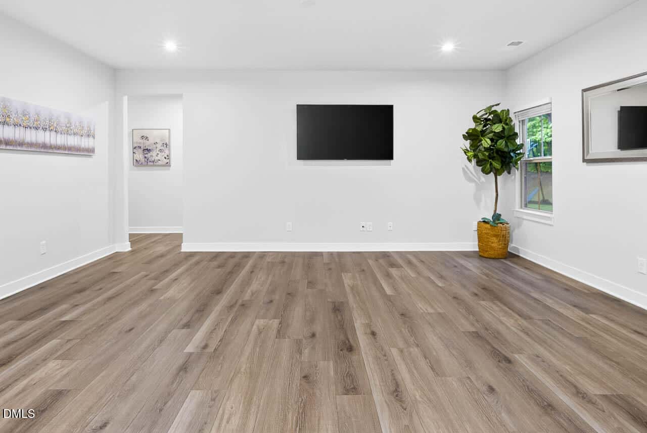 Spacious, modern room with wood flooring, white walls, a wall-mounted TV, a large potted plant by the window, a mirror, and wall art. Room is bright with recessed lighting and natural light.