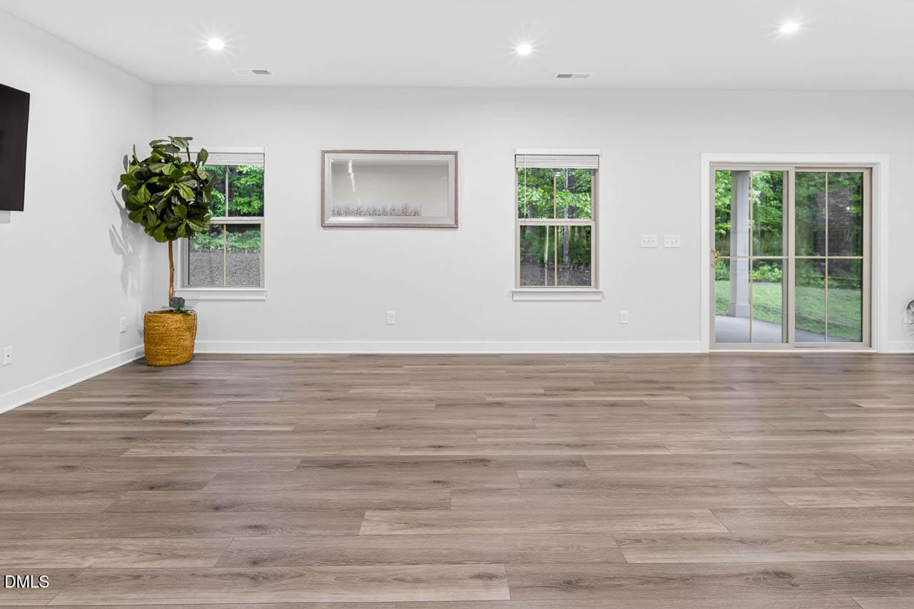 Spacious, empty room with light wood floors, white walls, two windows, a sliding glass door, a potted plant, and a wall-mounted TV on the left.