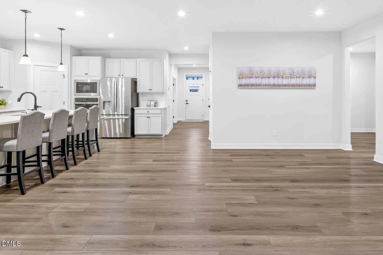 Modern kitchen with white cabinets, stainless steel appliances, a large island with four upholstered barstools, and light wood flooring. Open space leads to a hallway and features a wall-mounted artwork.