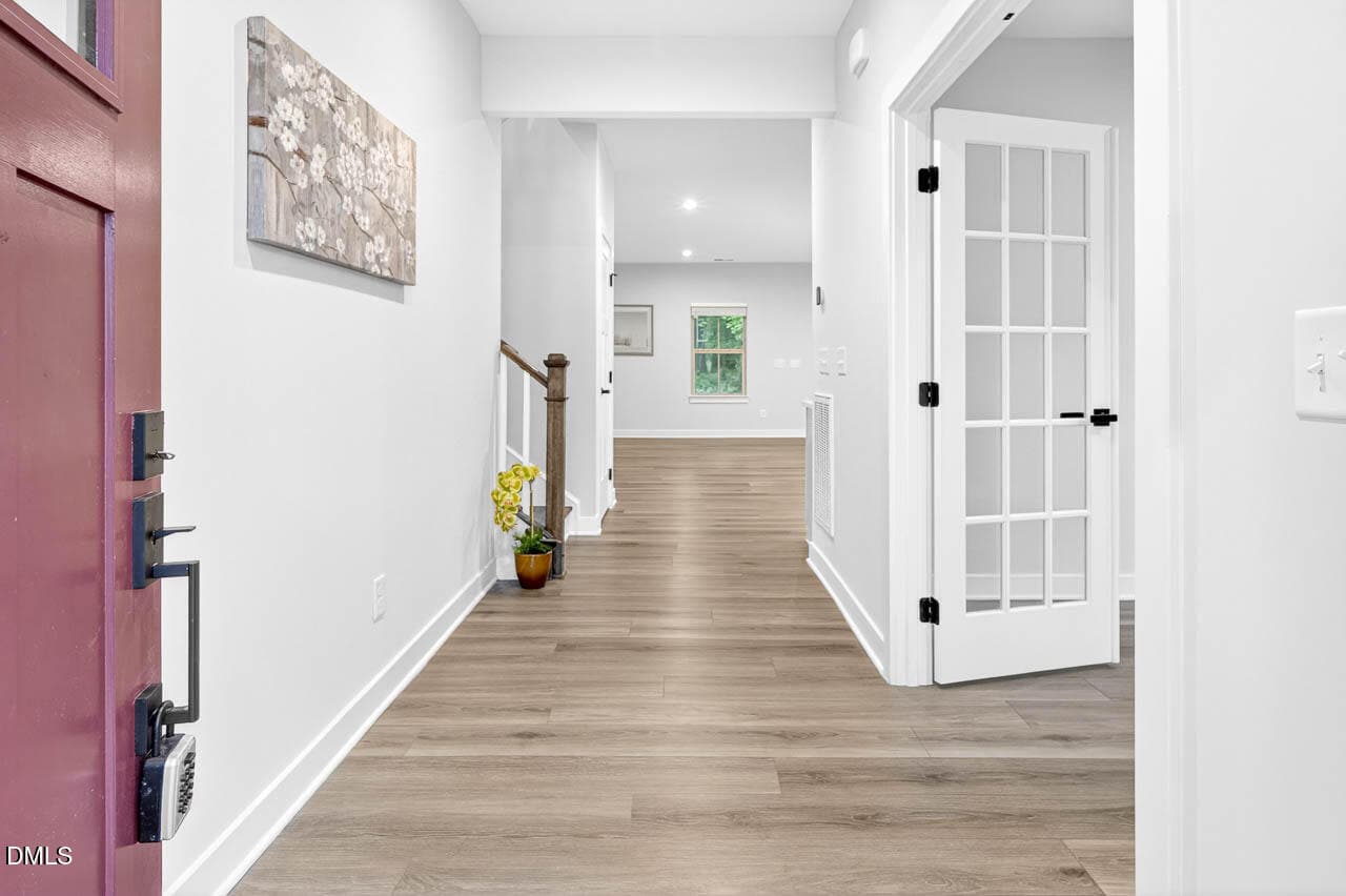 A bright, modern hallway with light wood floors, white walls, a glass-paneled door, stairs with a wooden banister, a potted plant, and a floral painting on the wall.