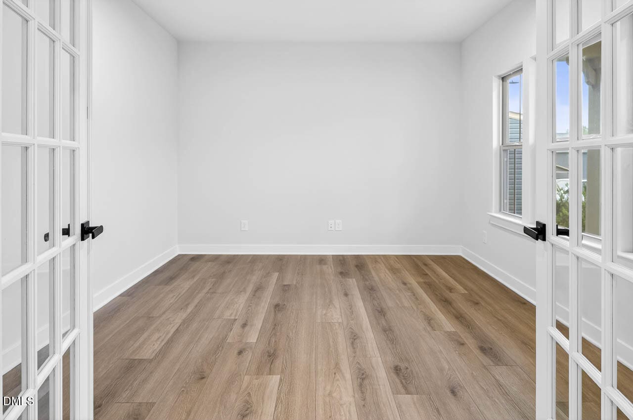 Empty room with light-colored walls, wide plank wood flooring, two windows on the right, and double French doors with glass panels partially open in the foreground.