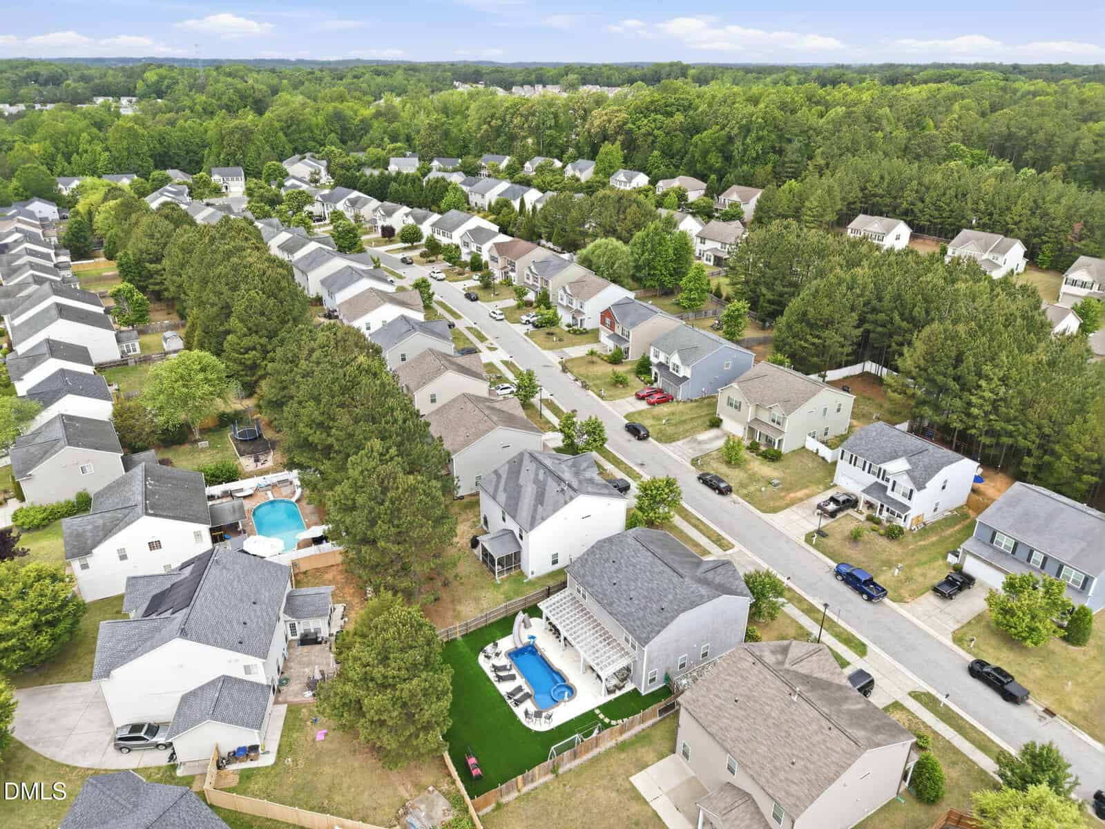 Aerial view of a suburban neighborhood with rows of houses, green lawns, trees, and a few backyards featuring swimming pools, under a partly cloudy sky.