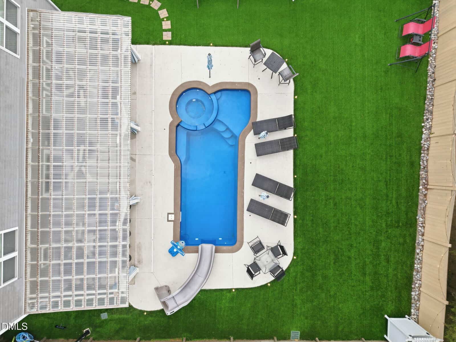 Aerial view of a backyard with a uniquely shaped swimming pool, surrounding lounge chairs, a slide, artificial grass, a covered patio on the left, and a red swing on the top right.