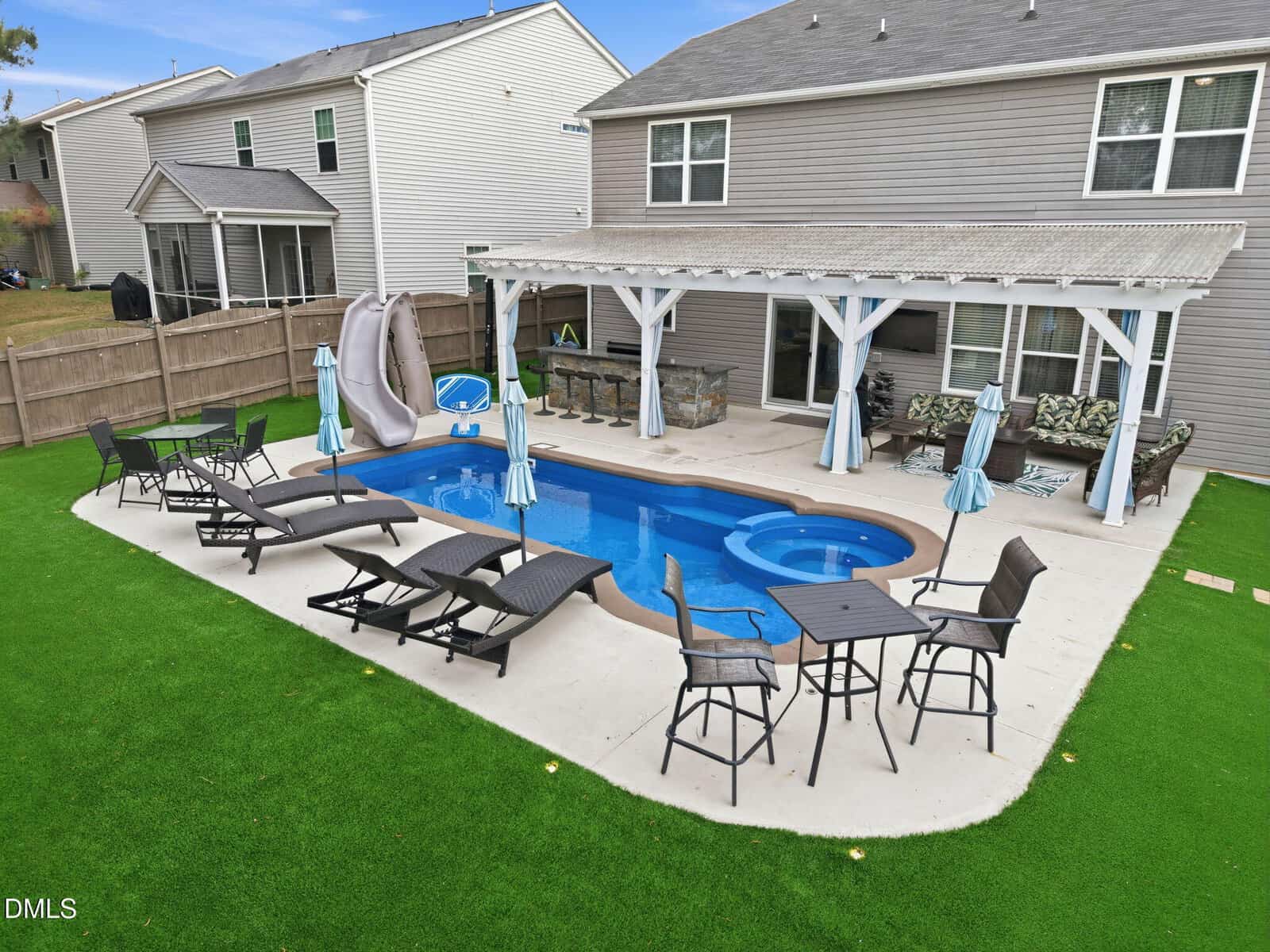 A backyard with a pool, spa, lounge chairs, tables, a slide, and a covered patio area with bar seating. The yard has artificial turf and a wooden fence, with two-story houses in the background.