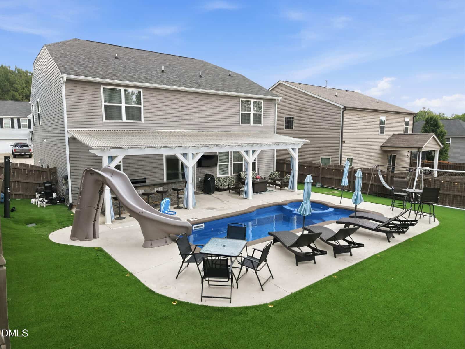 A backyard features a rectangular pool with a slide, lounge chairs, umbrellas, and a covered patio attached to a two-story house with beige siding and a fenced yard with artificial grass.