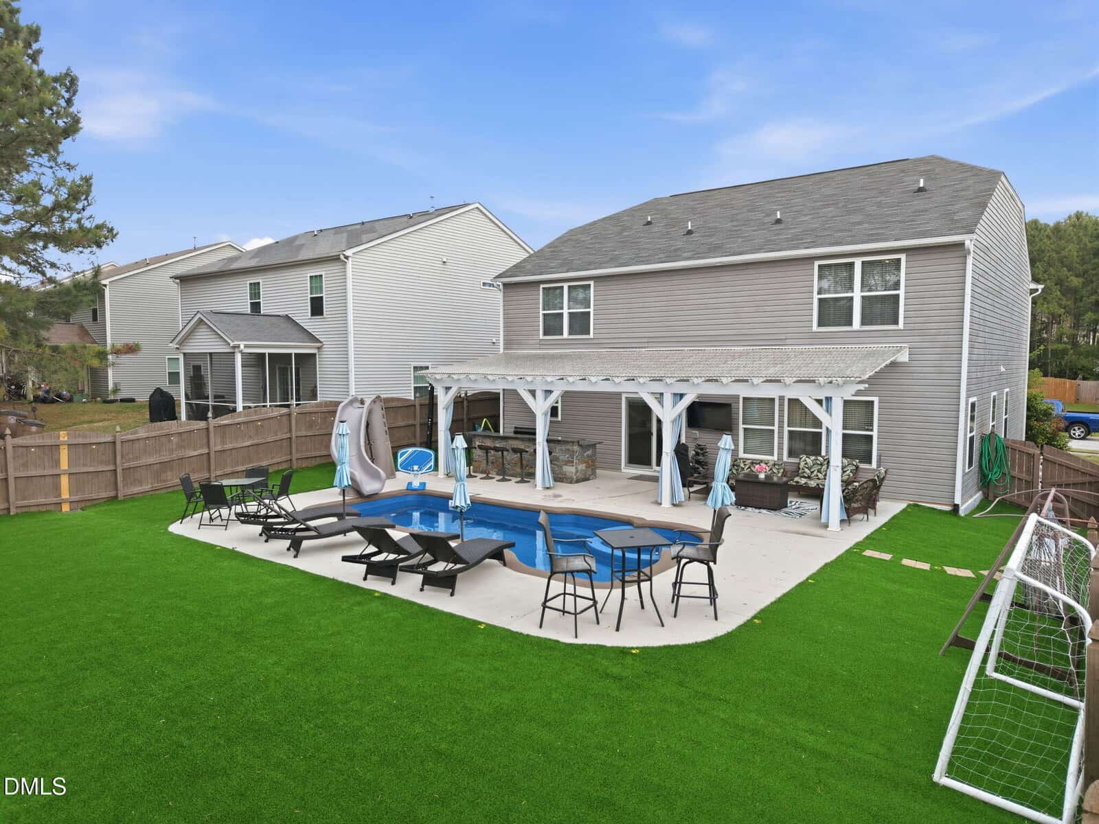 A backyard with artificial grass, a rectangular pool with a slide, lounge chairs, a pergola-covered sitting area, and a soccer goal, behind a two-story gray house.