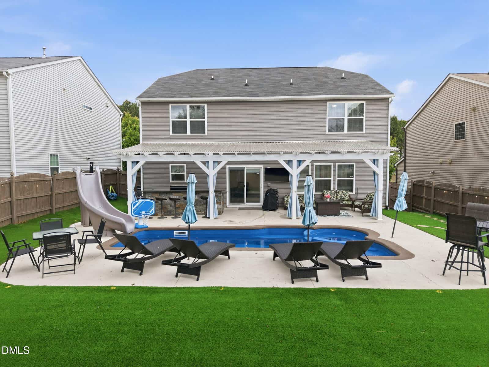 A backyard with a rectangular pool, waterslide, lounge chairs, umbrellas, and covered patio area attached to a two-story house, surrounded by artificial grass and a wooden fence.