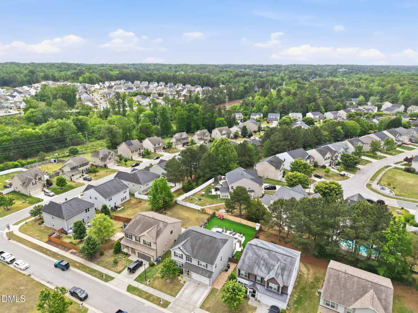 Aerial view of a suburban neighborhood with rows of houses, green lawns, trees, and winding streets, set against a backdrop of forested areas and a clear blue sky.