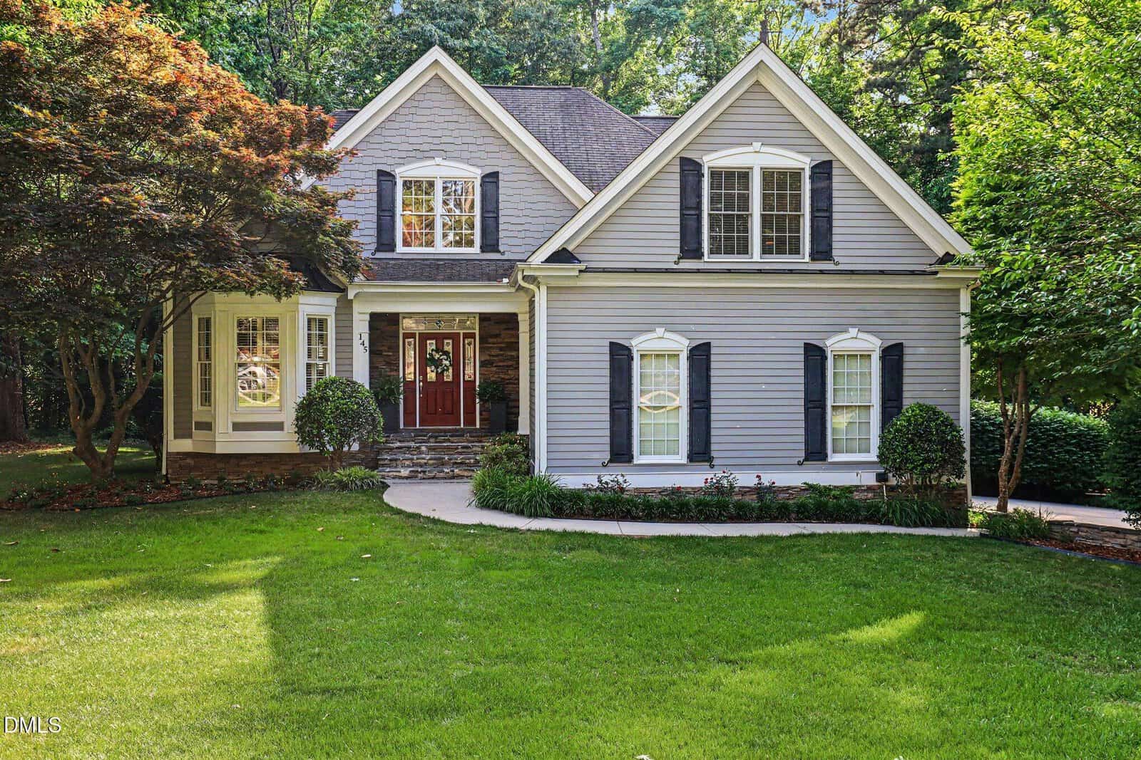A two-story gray house with white trim and dark shutters, a dark red front door, and a well-maintained lawn with trees and bushes, set in a suburban neighborhood.