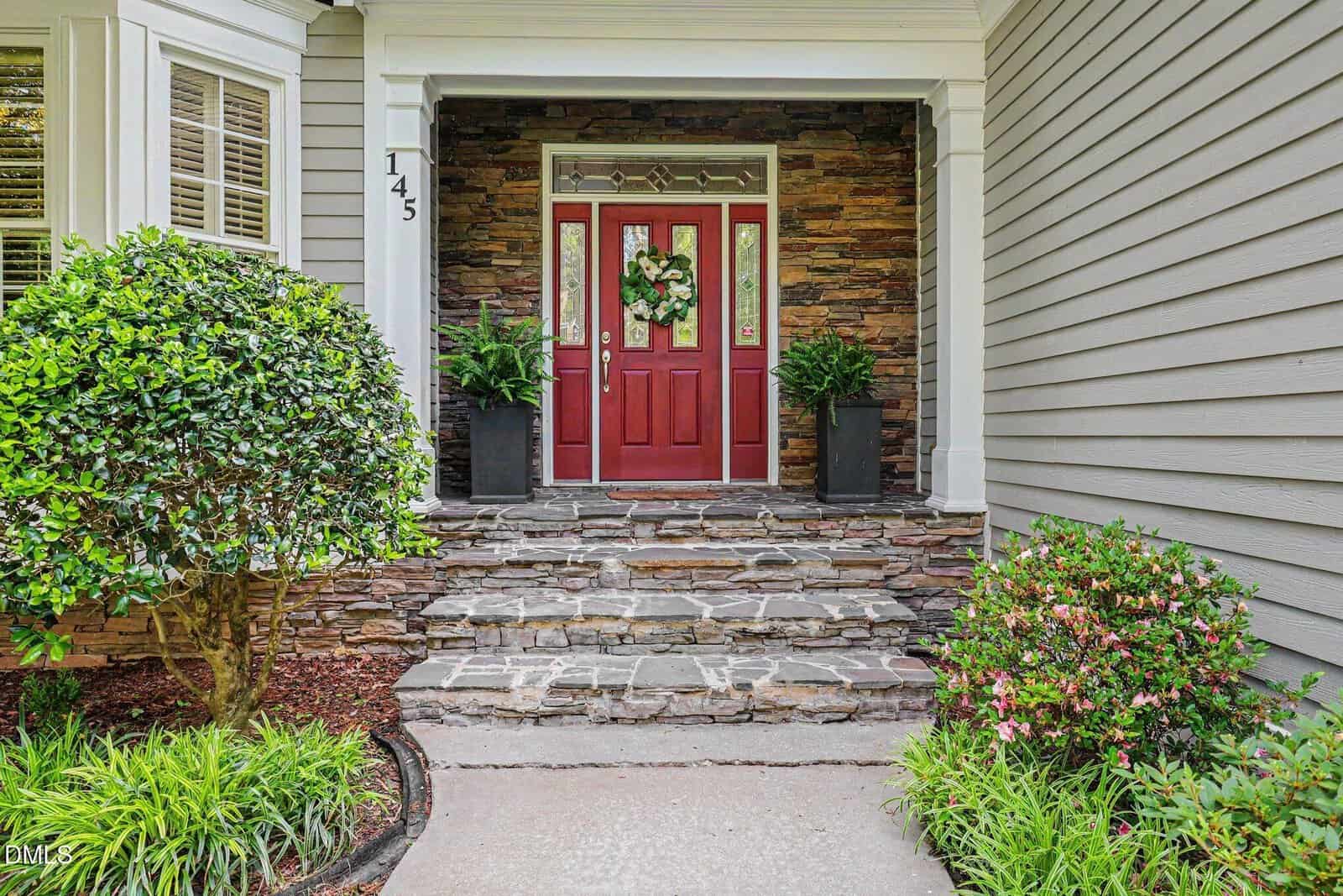 Front entrance of a house with a red door decorated with a wreath, flanked by two potted plants. Stone steps lead up to the door, surrounded by shrubs and beige siding. House number 145 is on a white column.