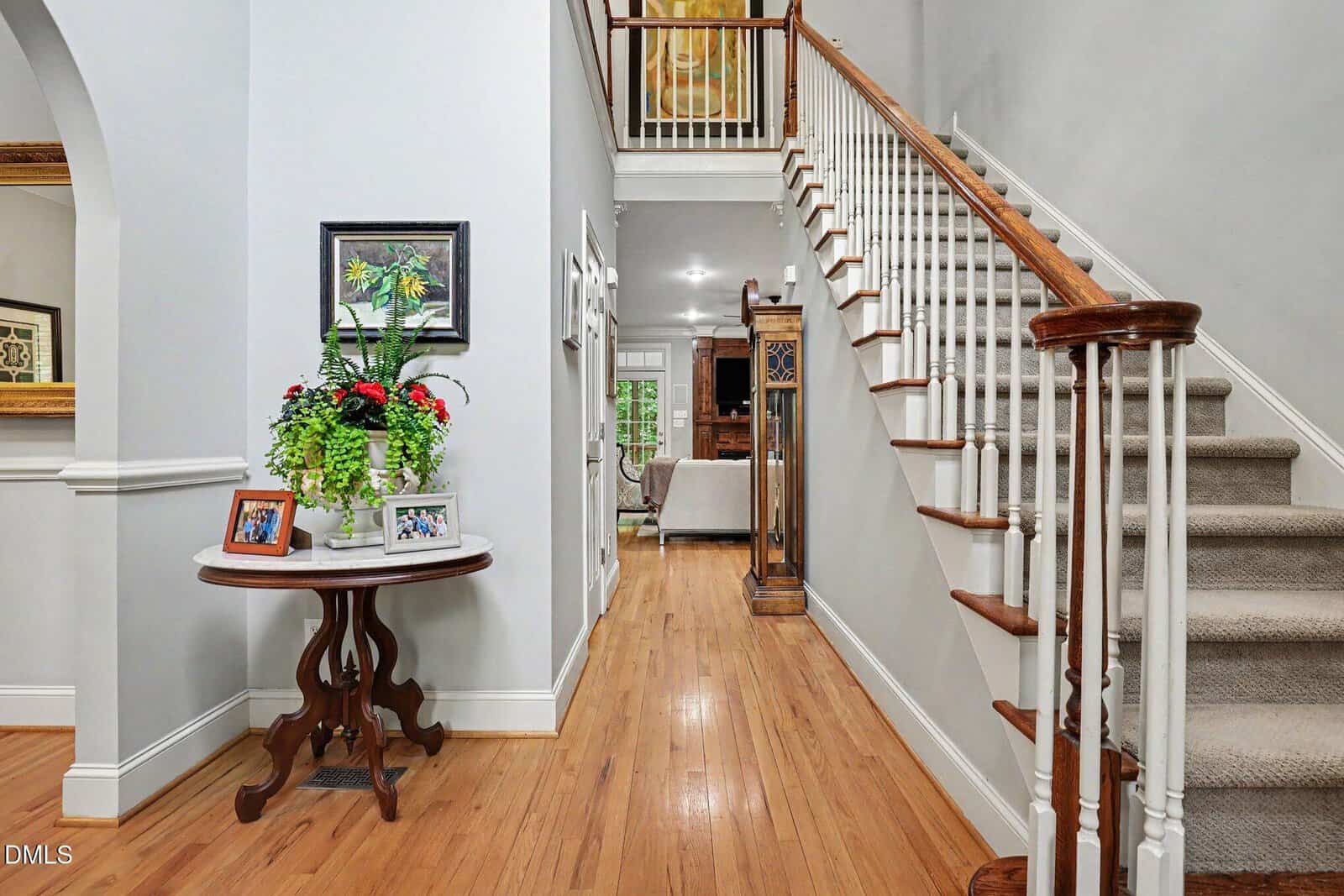 A bright hallway with wood floors and a carpeted staircase. A round table with plants and framed photos sits against the wall. The space leads into a living area with neutral decor.
