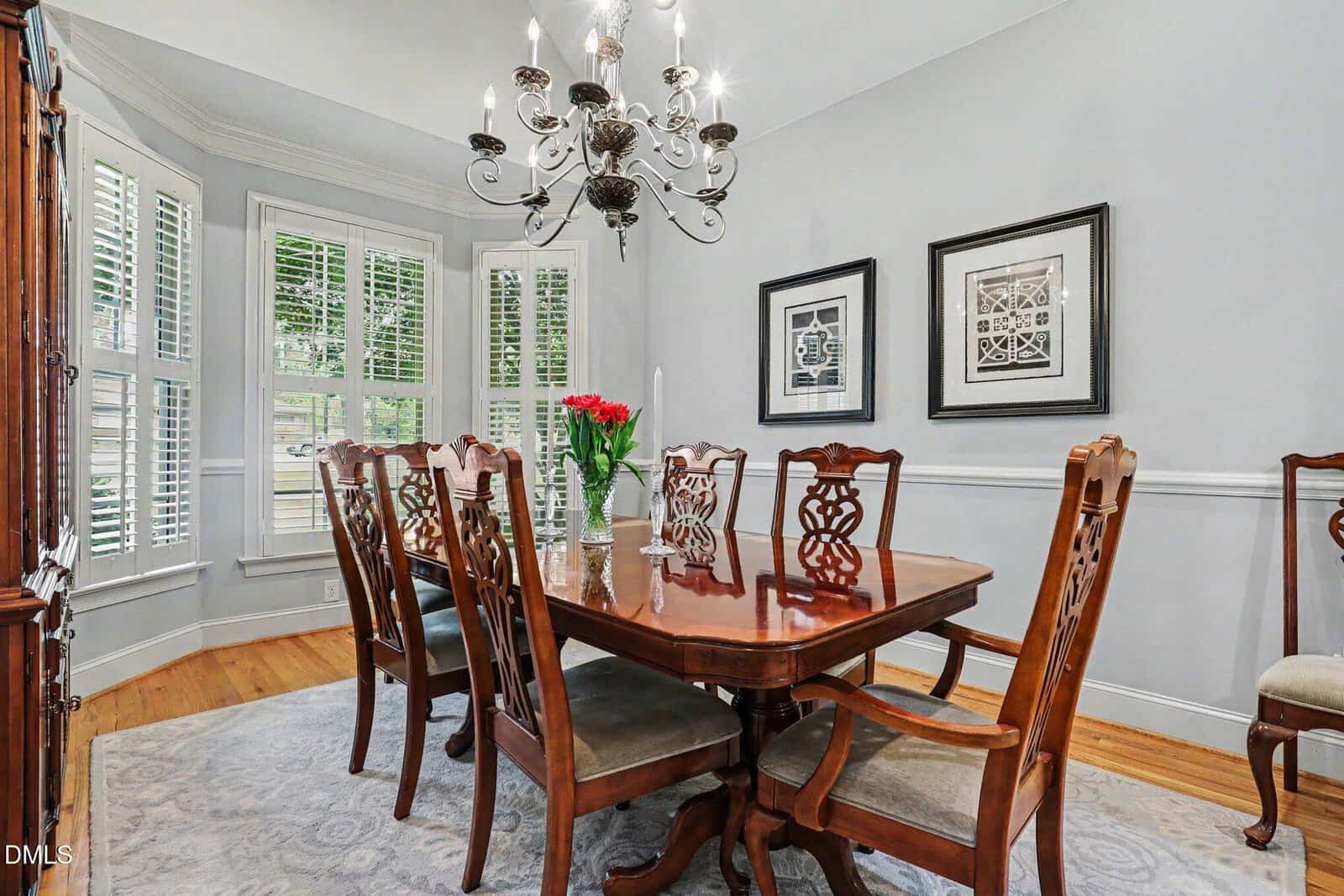 Elegant dining room with a wooden table and eight matching chairs, a chandelier overhead, light gray walls, two framed artworks, large windows with white shutters, and a vase of red flowers as the centerpiece.