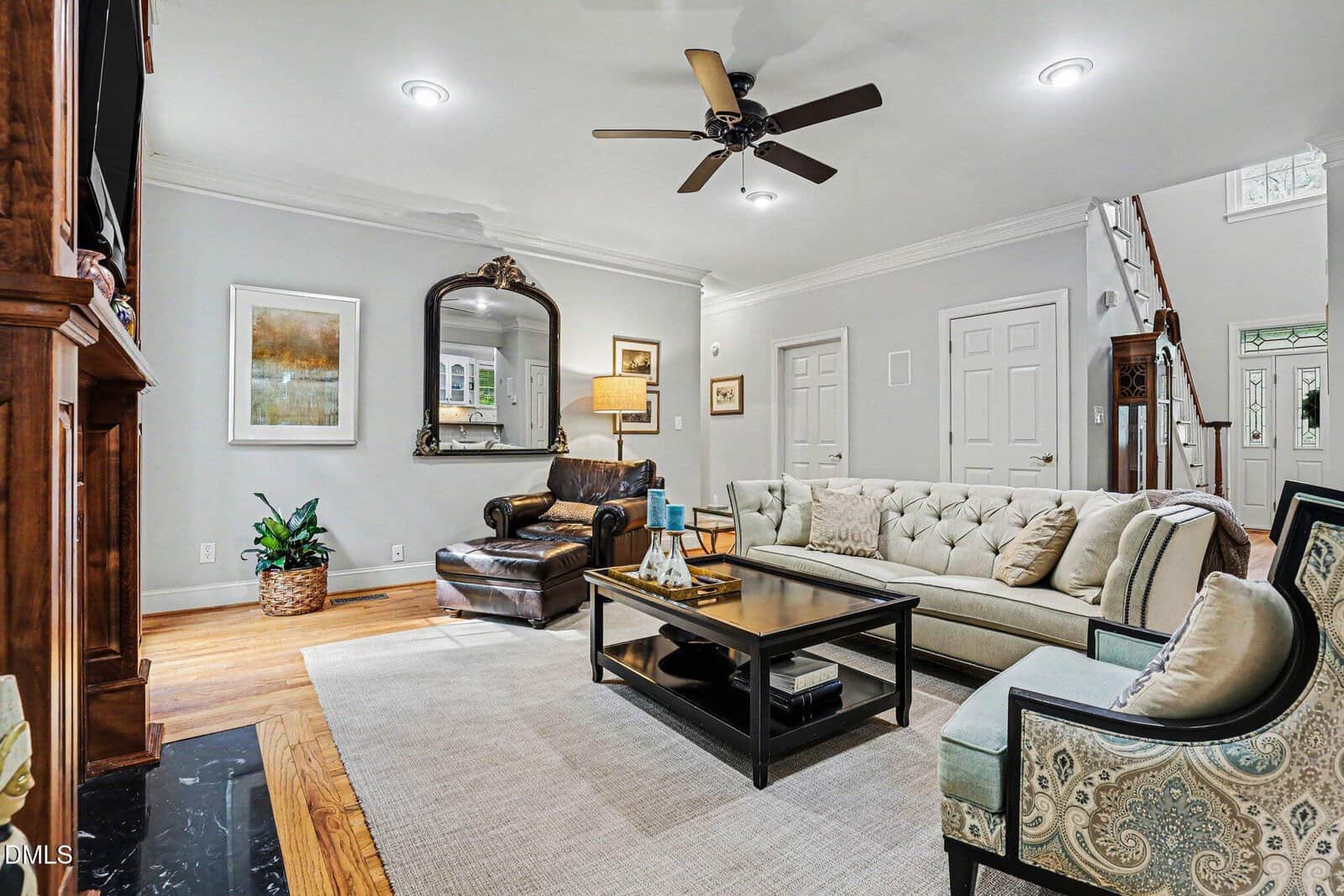 Elegant living room with a beige sofa, armchair, coffee table, large wall mirror, and framed art. Hardwood floors, neutral walls, ceiling fan, and a staircase are also visible. The space is well-lit and inviting.