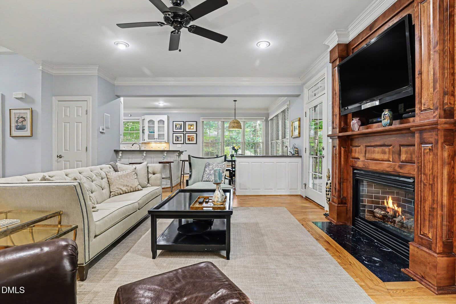 Elegant living room with a beige sofa, black coffee table, fireplace with a mounted TV, and large windows. Light wood floors, white walls, and ceiling fan create an inviting atmosphere with natural light.