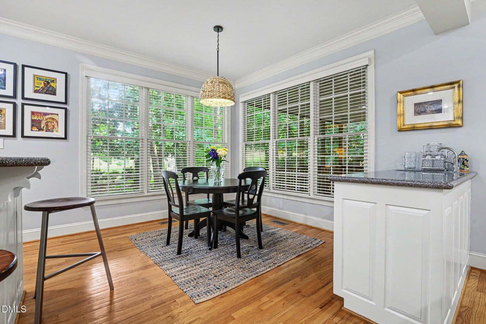 Bright breakfast nook with large windows, a round black dining table with four chairs, a woven pendant light, and a vase of flowers. White walls, wood floors, and bar stools complete the cozy space.