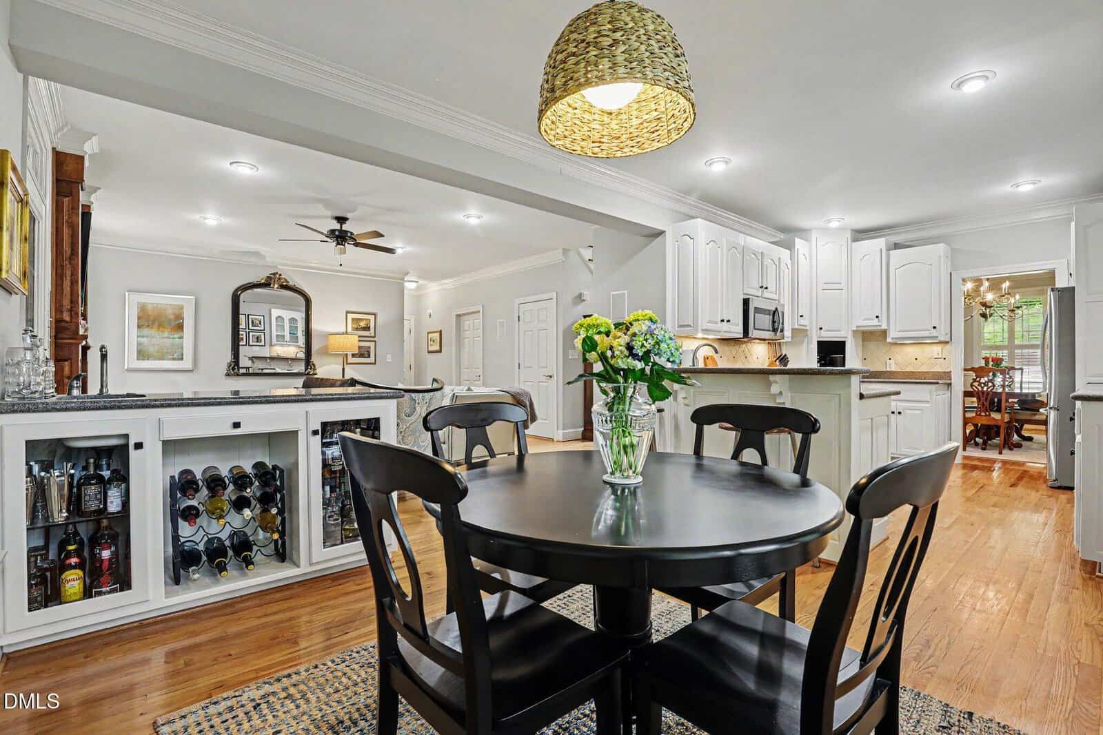 A modern kitchen and dining area with a black dining table, four chairs, a vase of flowers, white cabinets, a wine rack, wooden floors, and a hanging woven light fixture above the table.