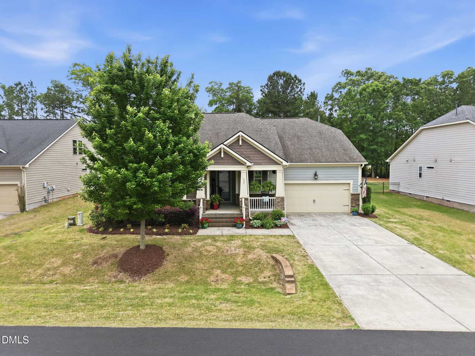 Single-story house with a front porch, attached two-car garage, and well-maintained lawn with a tree and shrubs, situated in a suburban neighborhood under a clear blue sky.