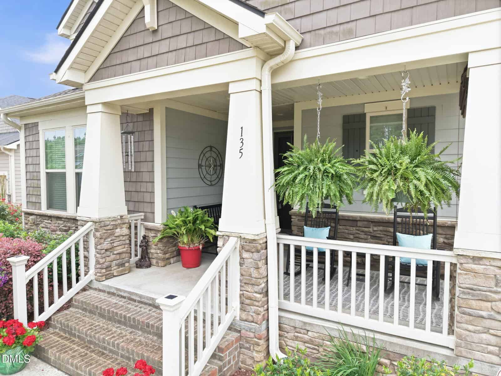 A charming front porch with stone columns, hanging ferns, two rocking chairs with blue pillows, a red potted plant, and a house number "123" on the pillar. Steps lead up to the porch, bordered by flowers and greenery.