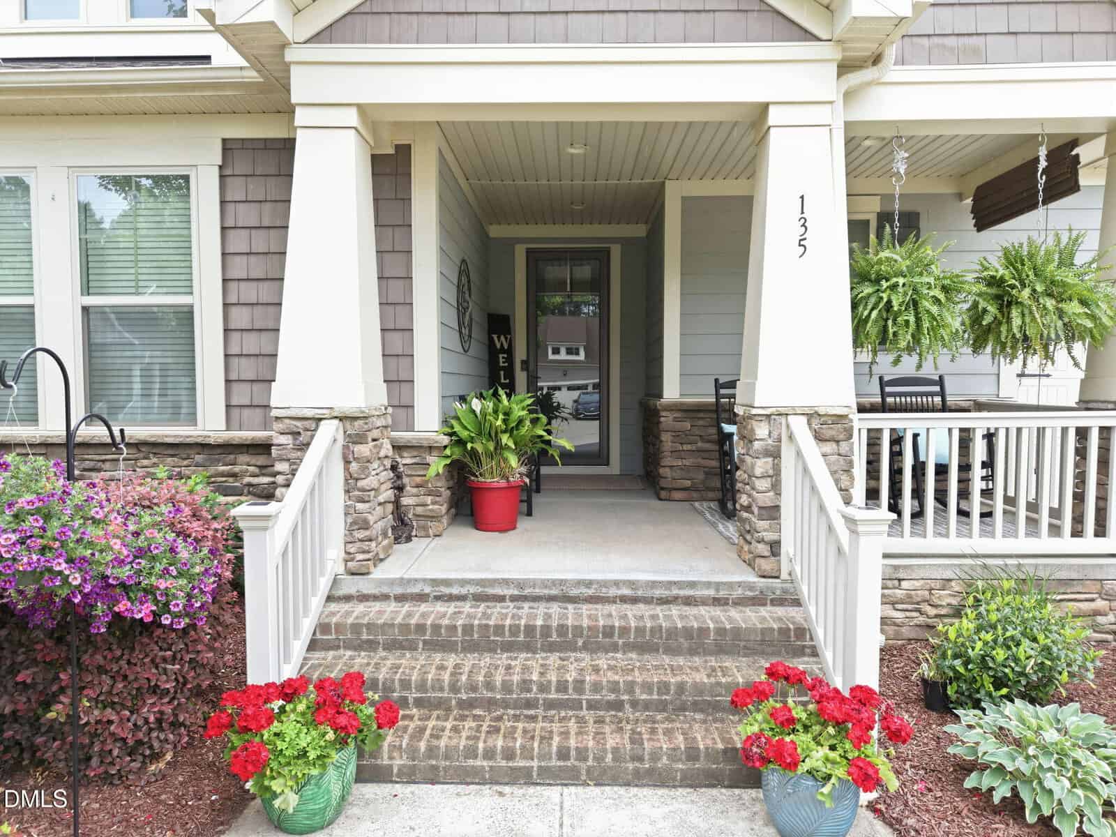 Front porch of a house with stone accents, potted red and purple flowers, large green ferns, a welcome sign by the door, and a covered sitting area with a porch swing and table. House number 125 is visible on the column.