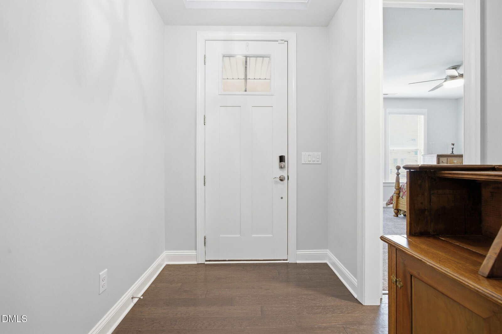 A white front door with a small window is set in a hallway with light gray walls and dark wood floors. To the right, a doorway leads to another room with a ceiling fan and window. A wooden cabinet is in the foreground.
