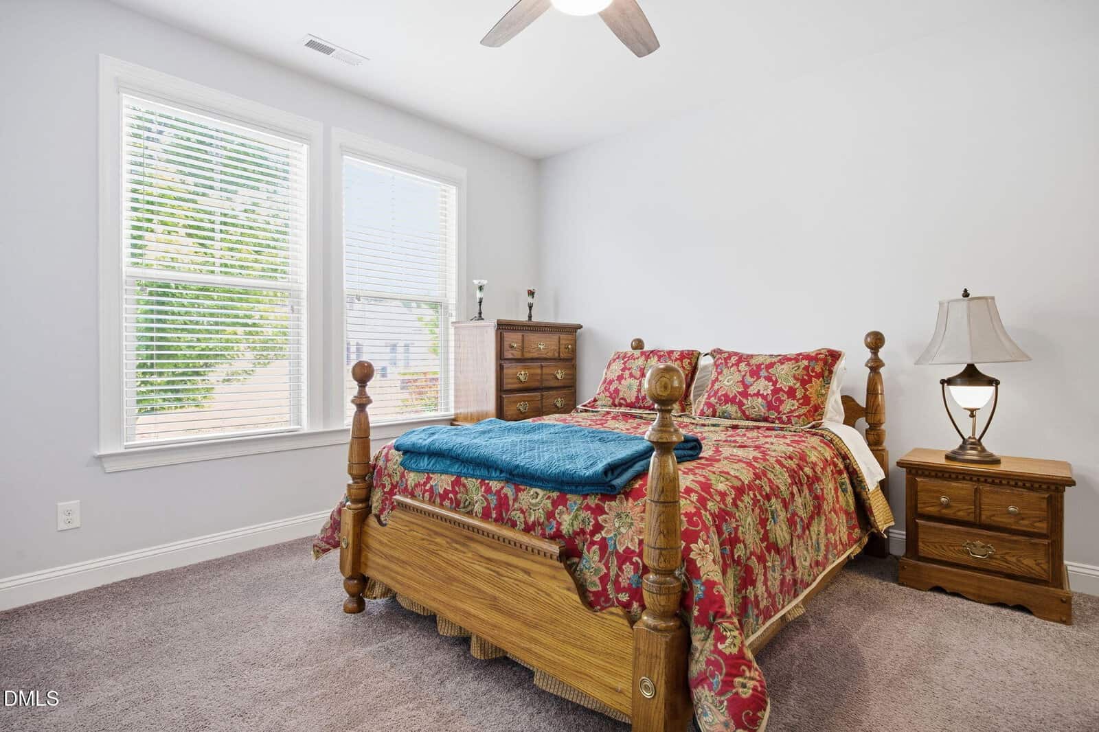 A bedroom with a wooden bed frame, floral bedding, blue blanket, two dressers, a lamp, and large windows with blinds letting in natural light.