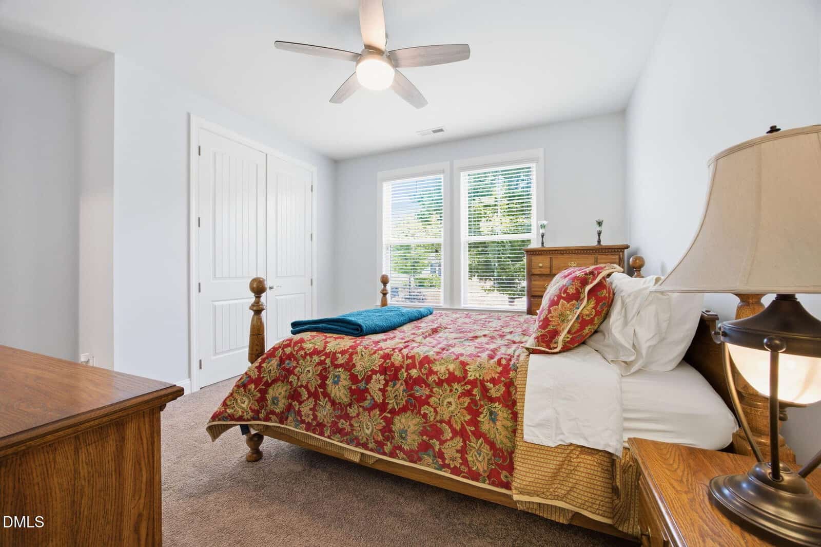 A bright bedroom with a wooden bed, colorful floral bedding, two wooden dressers, a nightstand with a lamp, ceiling fan, and large window with white blinds overlooking greenery.
