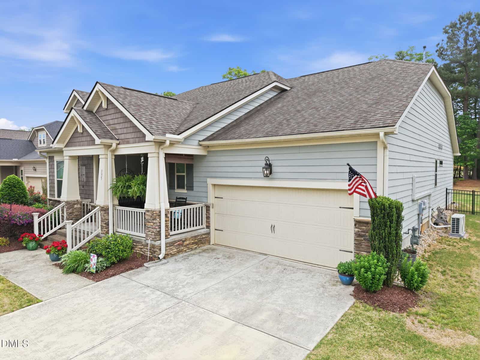 Single-story house with gray siding, stone accents, and a covered front porch. There is a two-car garage, an American flag, potted plants, and manicured landscaping in front of the home.
