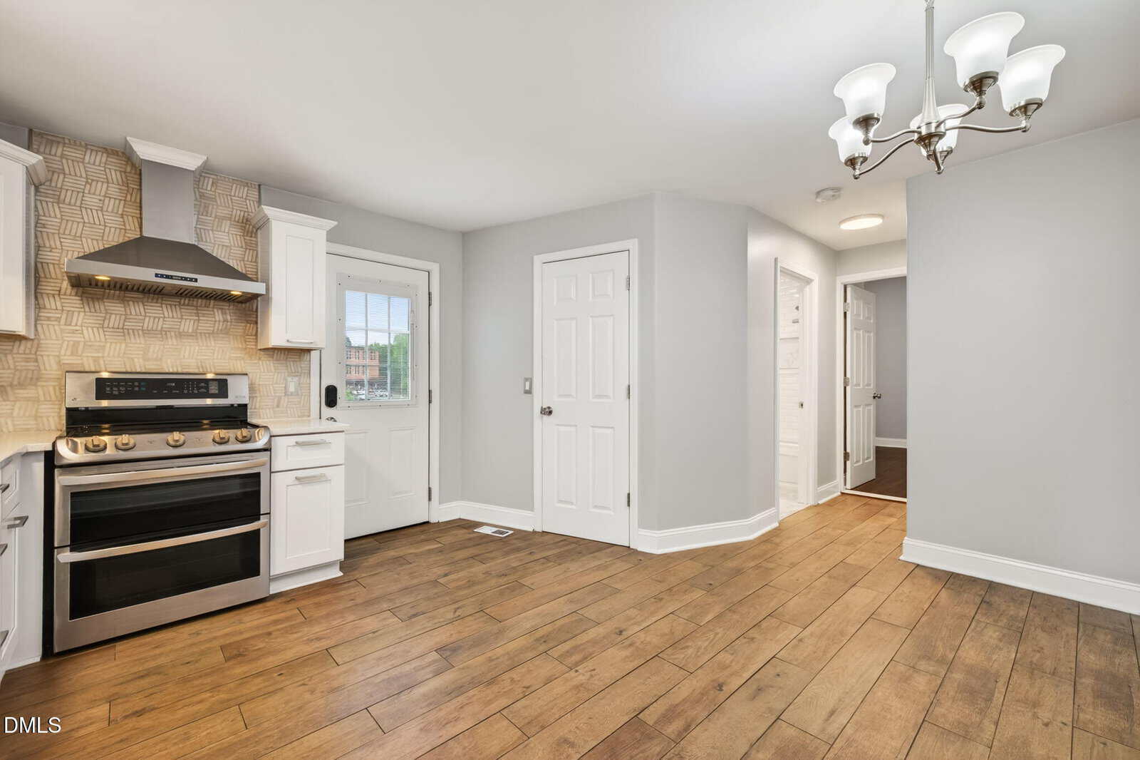 Modern kitchen with wood flooring, stainless steel stove and hood, white cabinets, patterned tile backsplash, a white door with window, two closed doors, and a chandelier. Hallway leads to other rooms.
