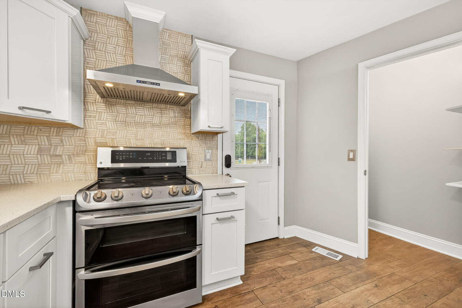 Modern kitchen with white cabinets, stainless steel stove and hood, patterned backsplash, light countertops, wood floor, and a door with a window leading outside. Open shelving is visible through the doorway on the right.