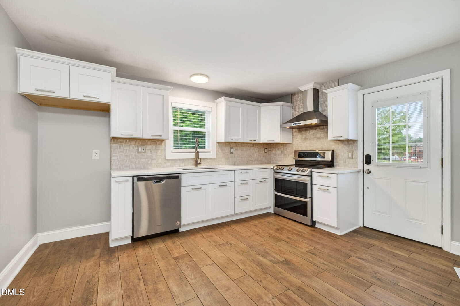 Bright kitchen with white cabinets, stainless steel appliances, light tile backsplash, wooden floor, window over sink, and a glass door leading outside. Walls are light gray, and there is ample counter space.