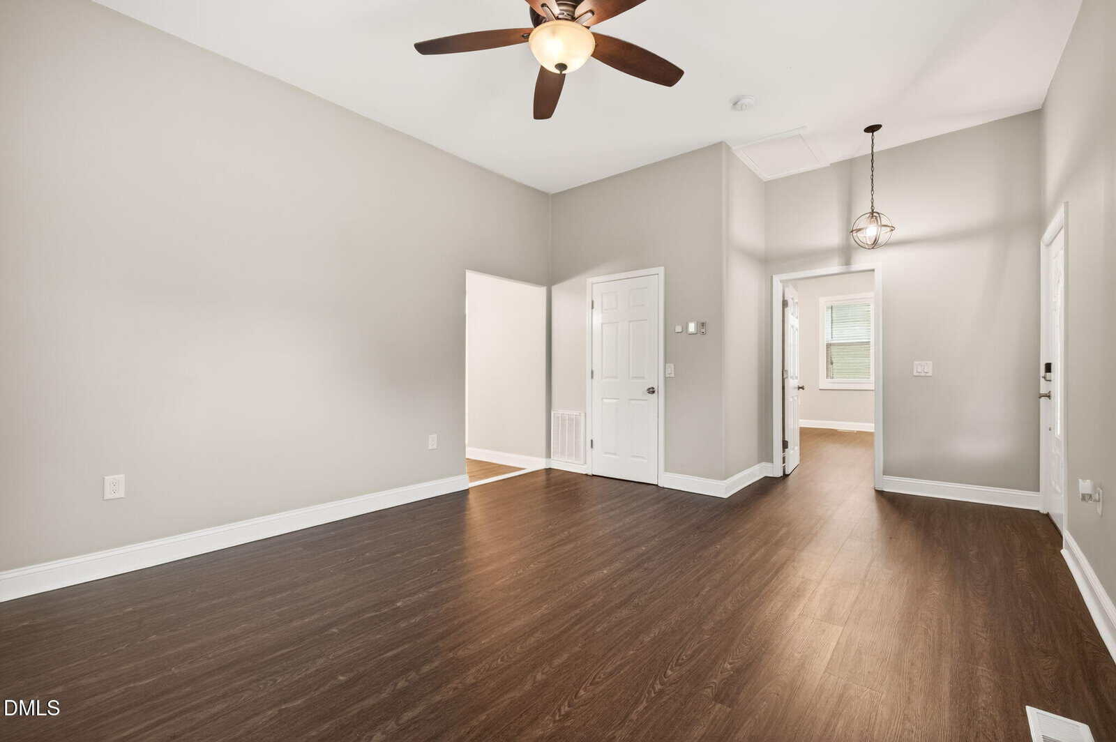 A spacious, empty room with light beige walls, dark wood flooring, a ceiling fan with lights, and a hallway leading to other rooms. Natural light enters through a window in the adjacent room.