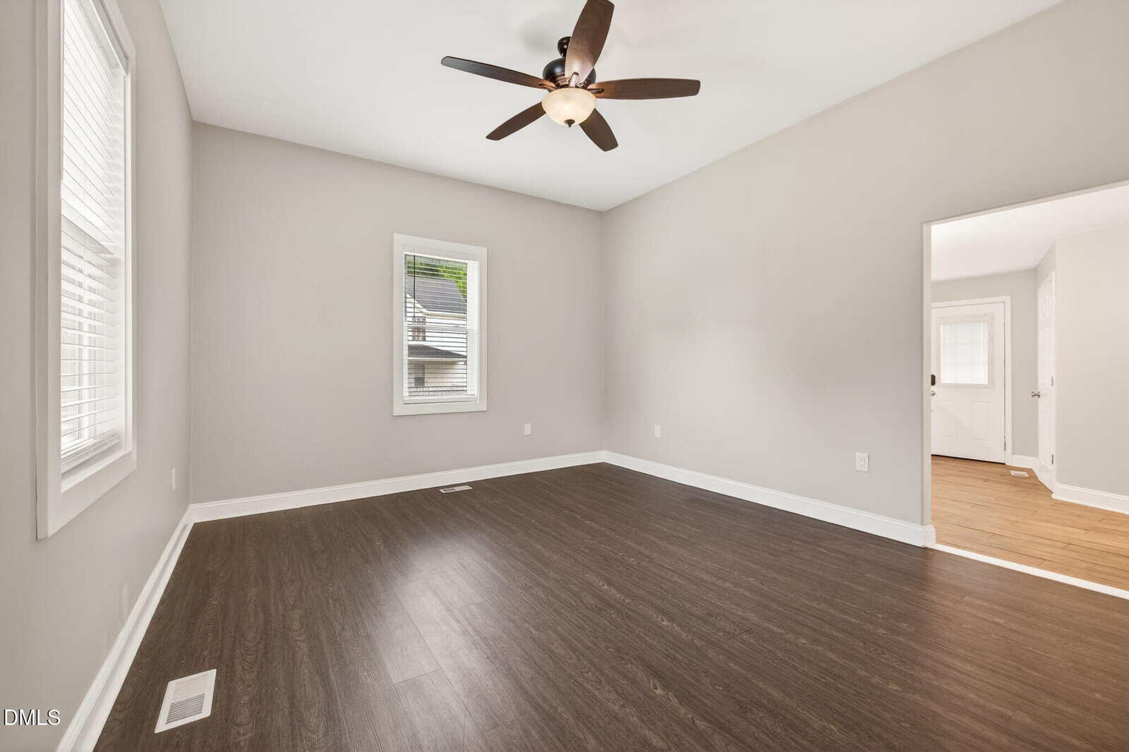 A spacious, empty room with light gray walls, dark wood flooring, a ceiling fan with lights, and two windows. An open doorway leads to another bright area with light-colored flooring.