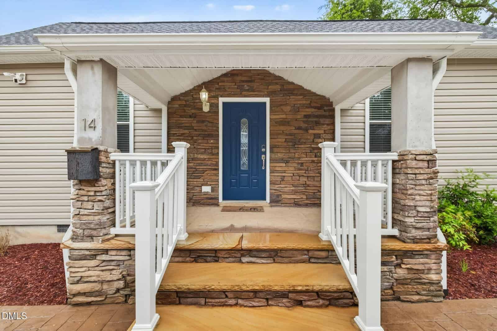 Front porch of a house with stone steps, white railings, tan siding, and a blue front door with a glass panel. The entryway is framed by decorative stonework and two white columns.