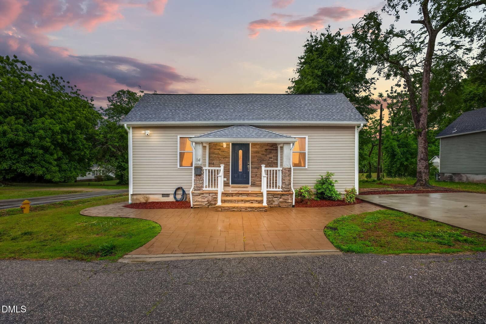 Small, light-colored house with a front porch, two columns, and yellow lights on either side of the door, surrounded by green trees and plants at sunset. Concrete driveway and walkway in front.