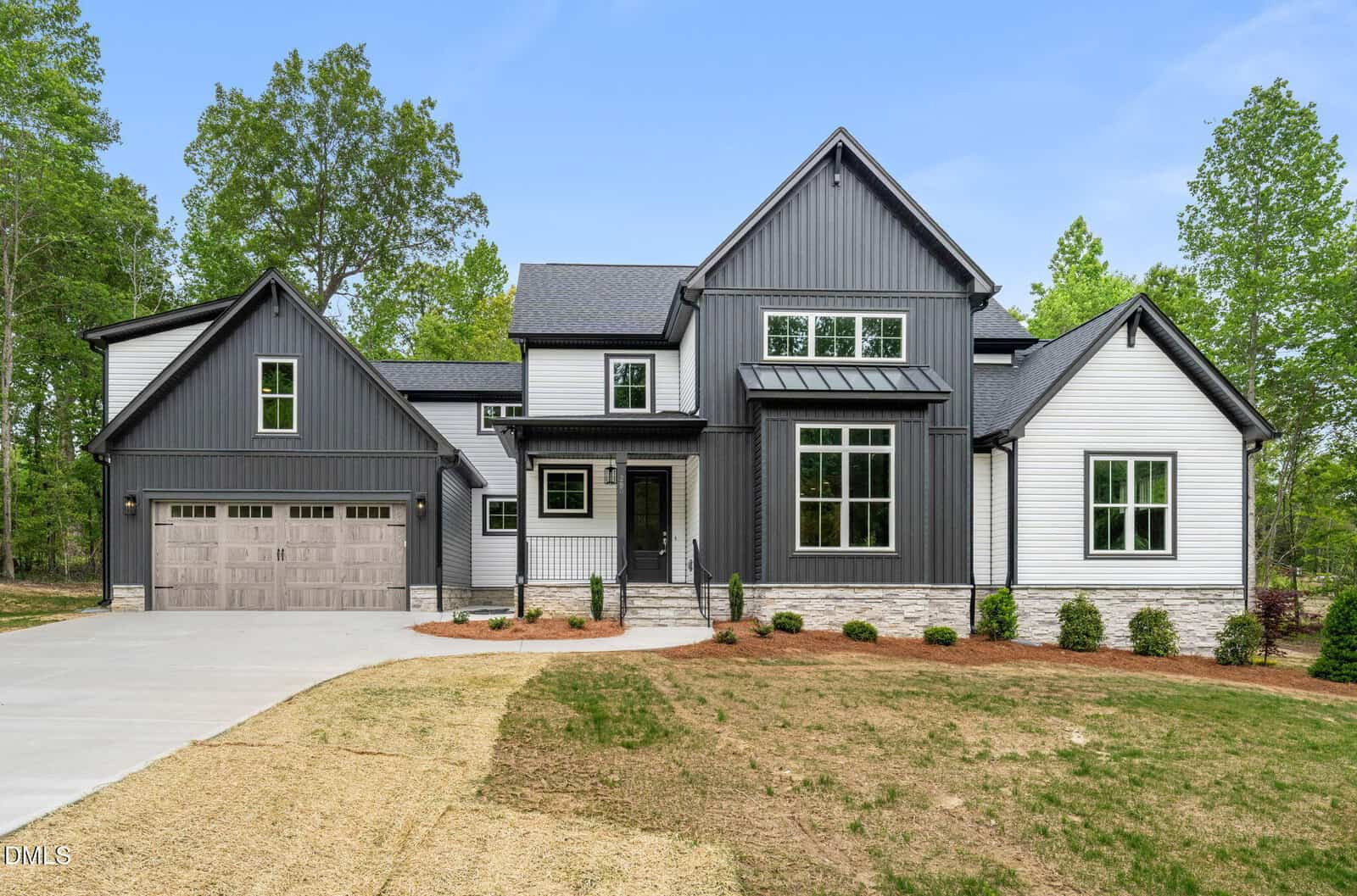 Modern two-story house with gray and white siding, large windows, a covered entryway, and a two-car garage. The home is surrounded by trees, with a partially grassed yard and a paved driveway.