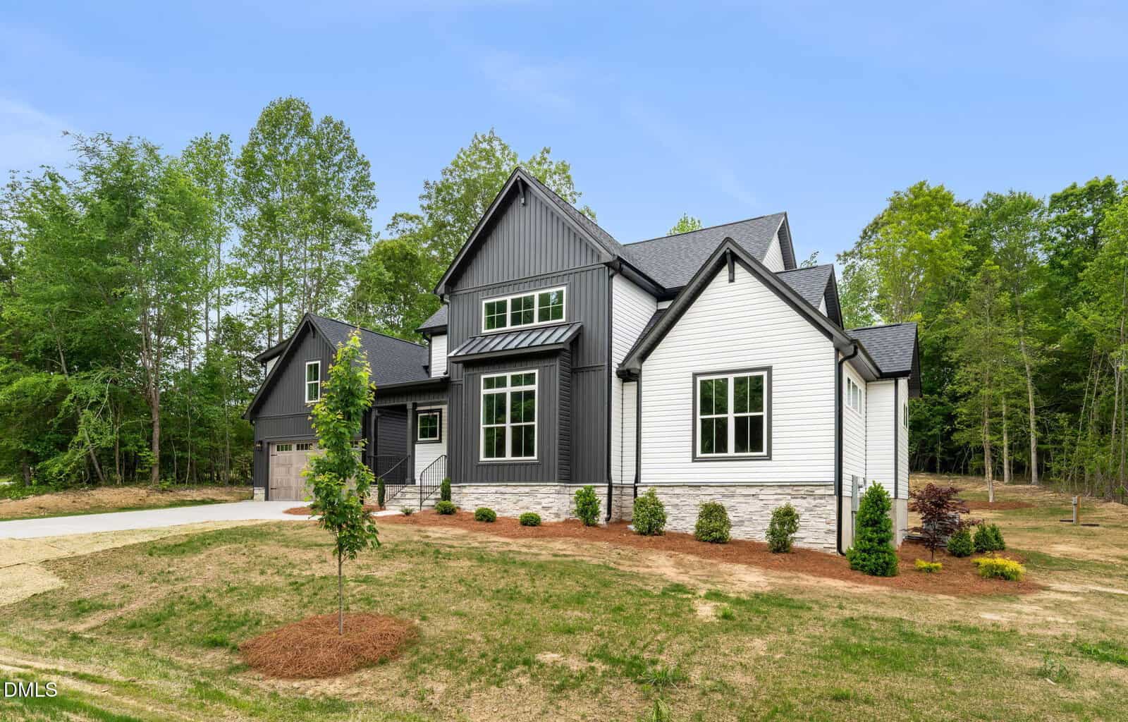 Modern two-story house with white and dark gray siding, large windows, and an attached garage. The yard has young trees, shrubs, and mulch, surrounded by grass and bordered by tall green trees.