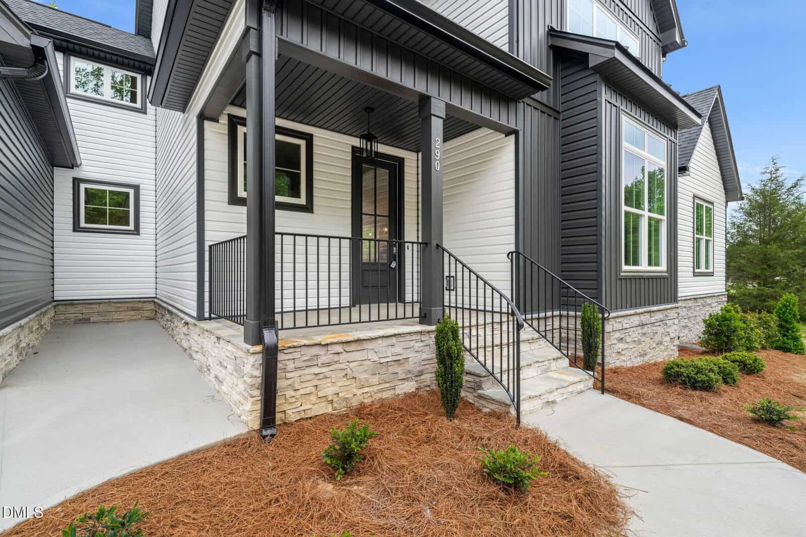 Modern house exterior with white and dark gray siding, stone foundation, black front door, porch with metal railing, and neatly landscaped shrubs and mulch. Concrete walkway leads to steps up to the entryway.