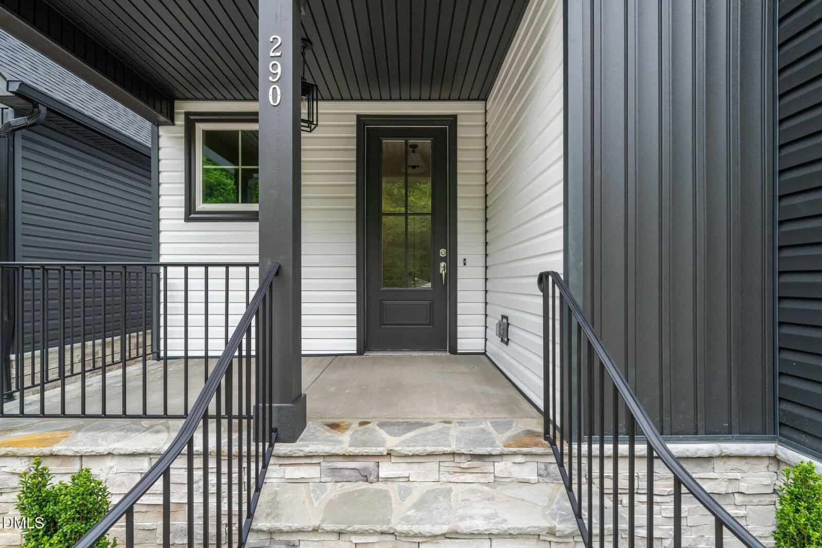 A modern house entrance with black railings, stone steps, white siding, and a black front door with glass panels. The porch is covered and features the house number 290 on a black column.
